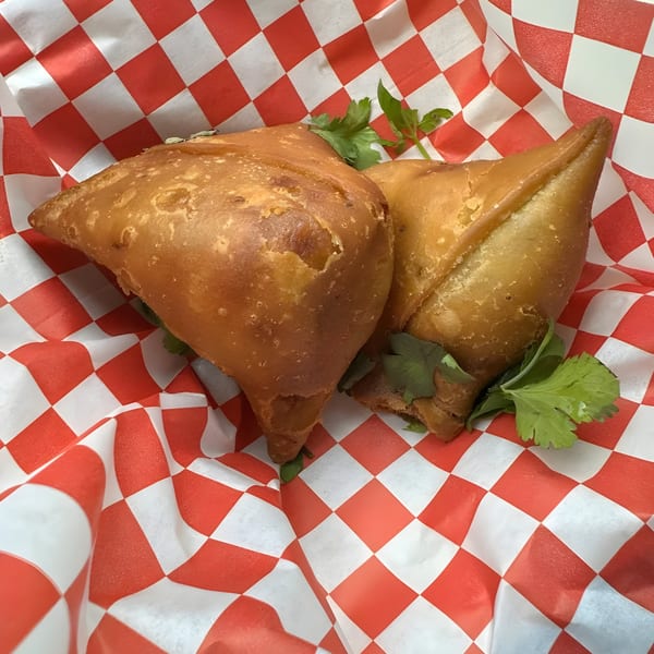 Two golden-brown samosas with cilantro, served in a red and white checkered paper basket.