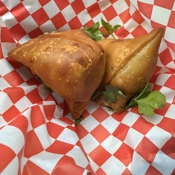 Two golden-brown samosas with cilantro garnish, served in a red and white checkered paper basket.
