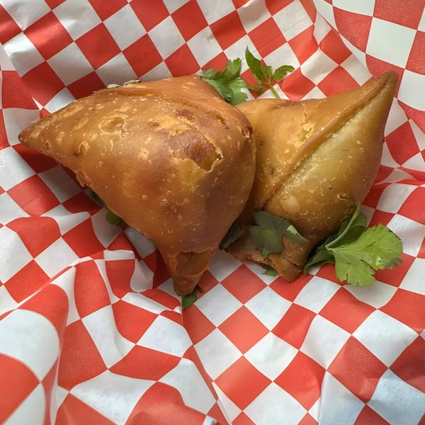 Two golden-brown samosas with cilantro garnish, served on red and white checkered paper.